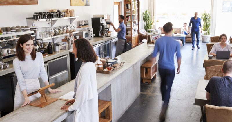 People in a cozy cafe, some engaged with laptops, perhaps working on social media strategies for small businesses. A barista assists a customer at the counter while another person walks by, and others are seated at tables in the background, enjoying the lively ambiance.